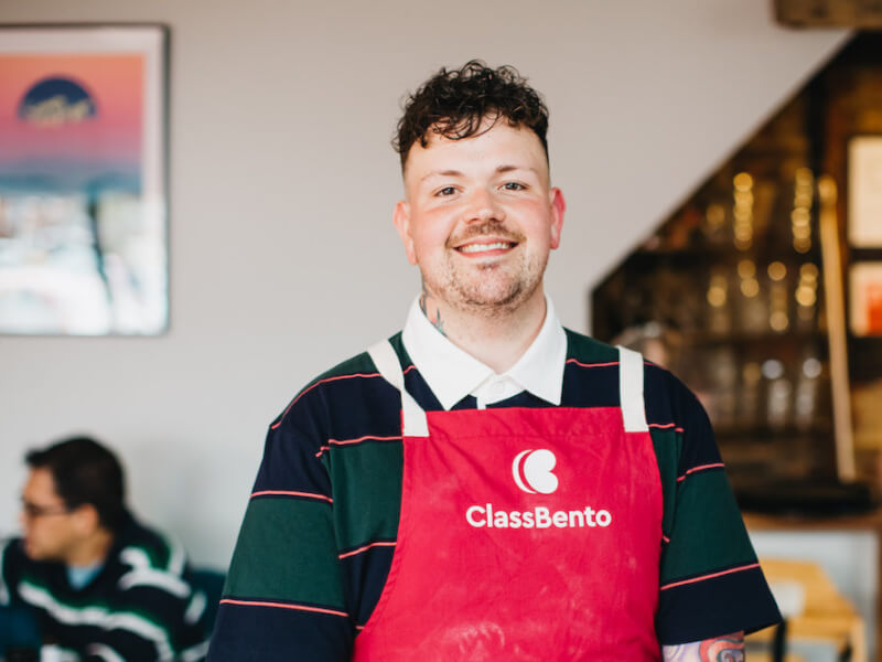 A man in a red ClassBento apron relaxing and smiling at a workshop